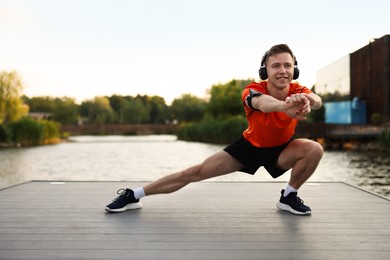 Photo of Smiling sportsman in headphones and armband doing morning exercise on pier near river