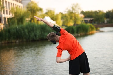 Photo of Sportsman in headphones and armband doing morning exercise near river, back view