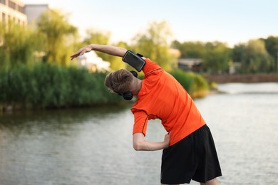 Photo of Sportsman in headphones and armband doing morning exercise near river, back view