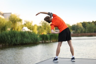 Photo of Sportsman in headphones and armband doing morning exercise on pier near river, back view. Space for text