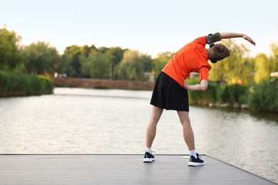 Photo of Sportsman in headphones and armband doing morning exercise on pier near river, back view. Space for text