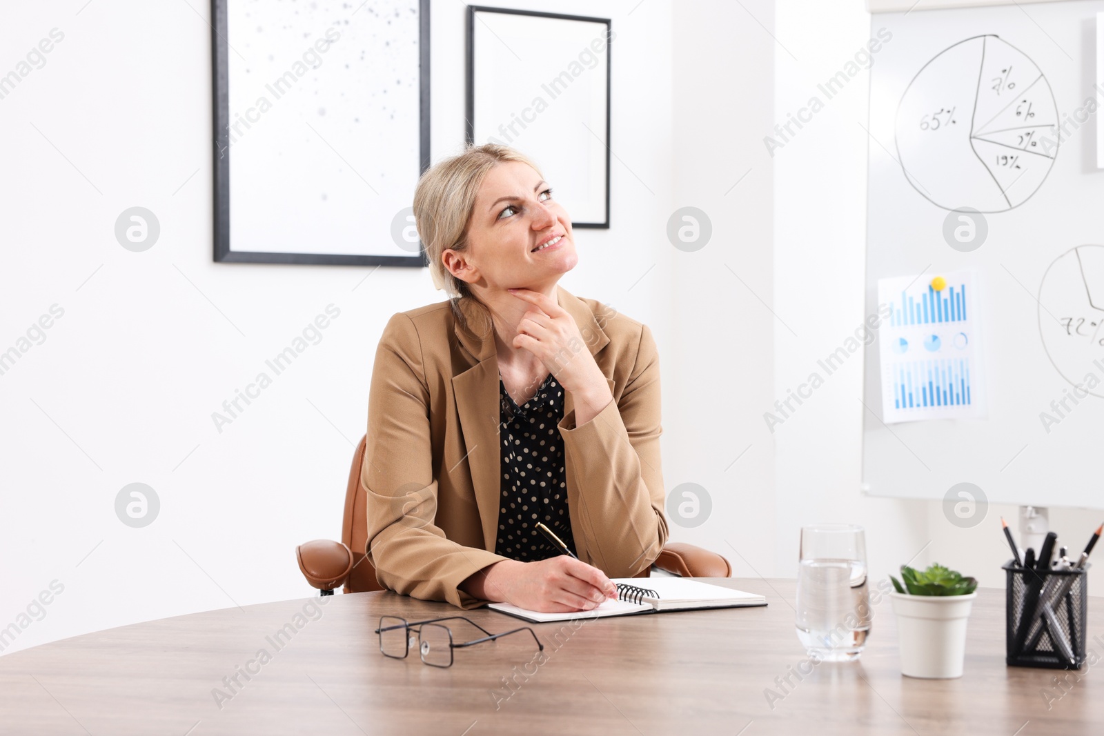 Dreams come true. Happy woman thinking about something while taking notes at wooden table indoors Photo of Dreams come true. Happy woman thinking about something while taking notes at wooden table indoors