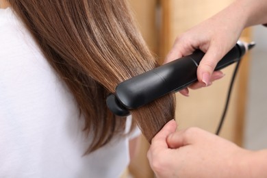 Photo of Hairdresser straightening woman's hair with flat iron in salon, closeup