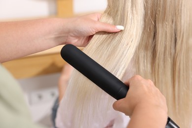 Photo of Hairdresser straightening woman's hair with flat iron in salon, closeup