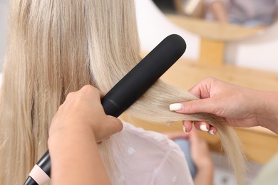 Photo of Hairdresser straightening woman's hair with flat iron in salon, closeup