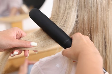 Photo of Hairdresser straightening woman's hair with flat iron in salon, closeup