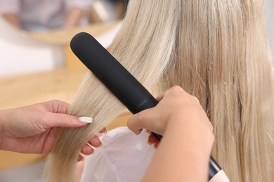 Photo of Hairdresser straightening woman's hair with flat iron in salon, closeup