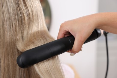 Photo of Hairdresser straightening woman's hair with flat iron in salon, closeup