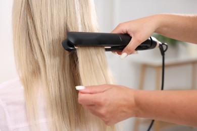 Photo of Hairdresser straightening woman's hair with flat iron in salon, closeup