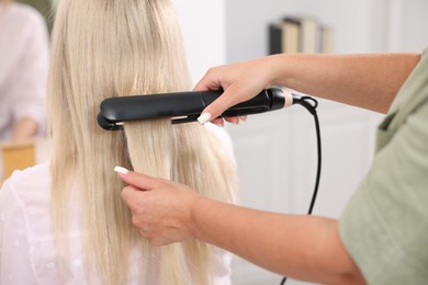 Photo of Hairdresser straightening woman's hair with flat iron in salon, closeup