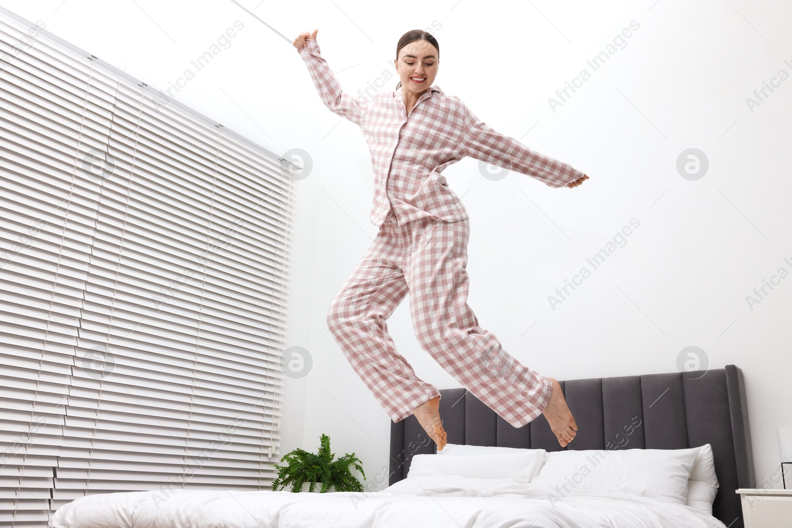 Happy woman in pyjama jumping on bed at home, low angle view Photo of Happy woman in pyjama jumping on bed at home, low angle view