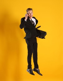 Photo of Businessman with briefcase jumping while talking on smartphone against orange background