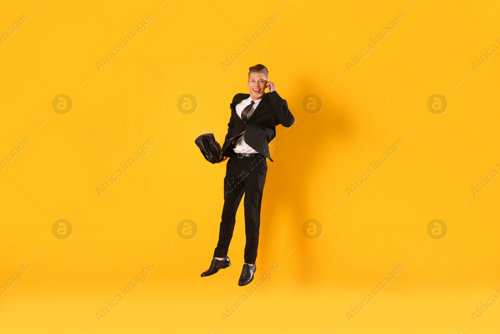 Businessman with briefcase jumping while talking on smartphone against orange background Photo of Businessman with briefcase jumping while talking on smartphone against orange background