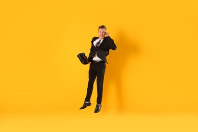 Photo of Businessman with briefcase jumping while talking on smartphone against orange background