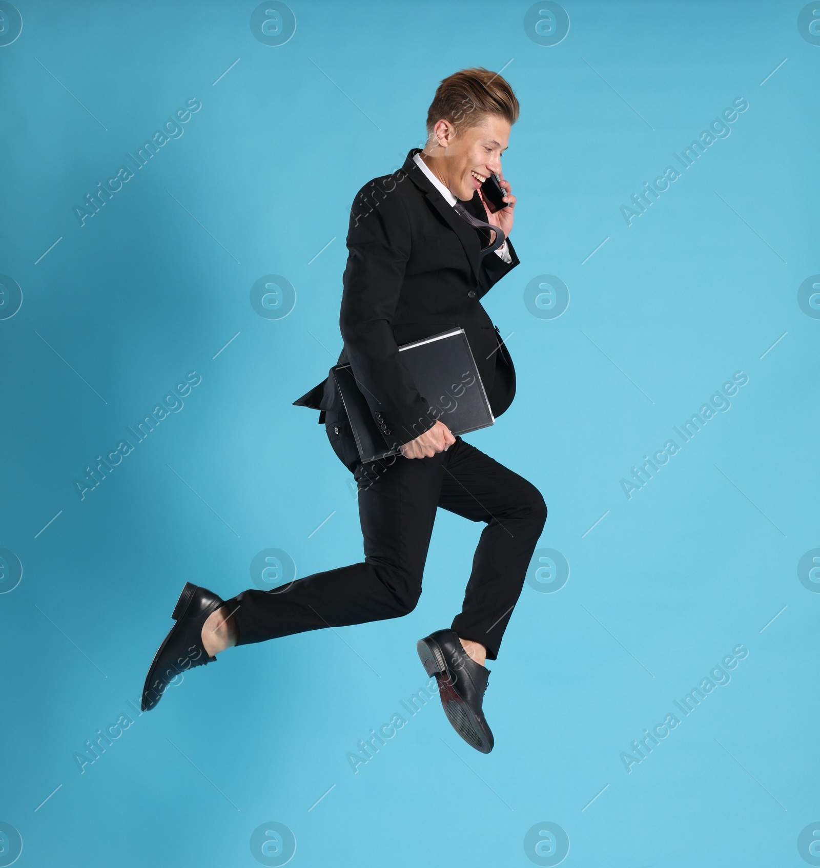 Businessman with folder jumping while talking on smartphone against light blue background Photo of Businessman with folder jumping while talking on smartphone against light blue background