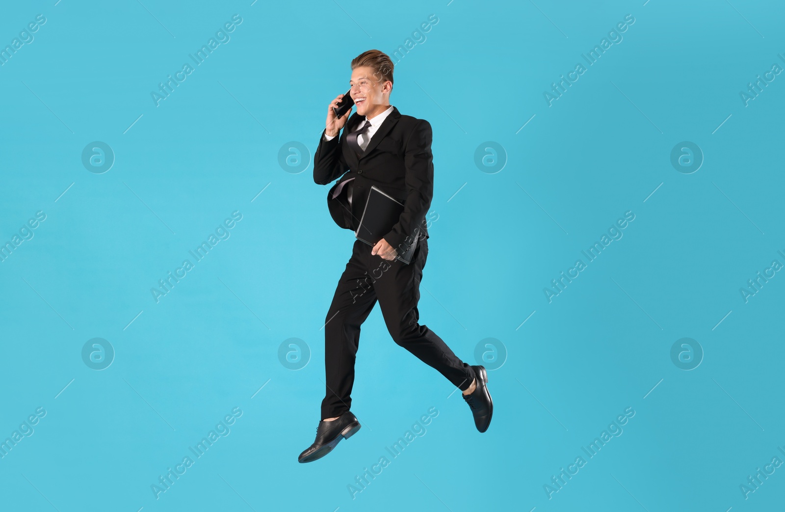 Businessman with folder jumping while talking on smartphone against light blue background Photo of Businessman with folder jumping while talking on smartphone against light blue background