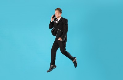 Photo of Businessman with folder jumping while talking on smartphone against light blue background