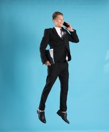 Photo of Businessman with folder jumping while talking on smartphone against light blue background