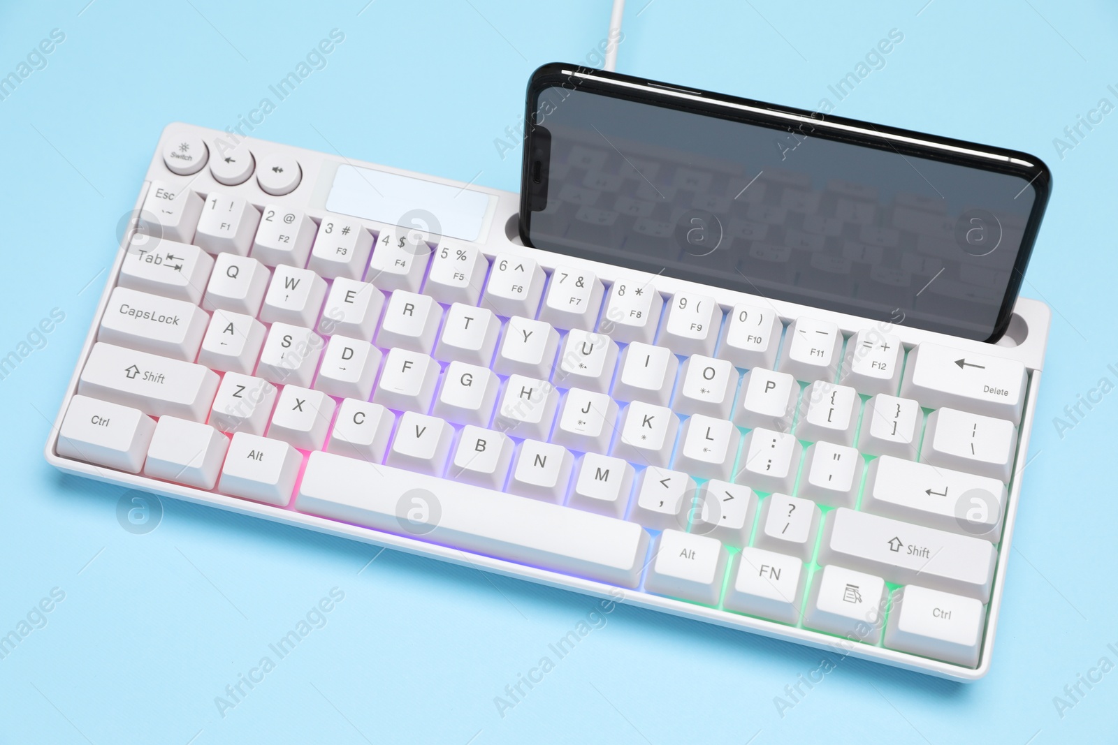 Photo of Modern white keyboard with RGB lighting and smartphone on light blue background, above view