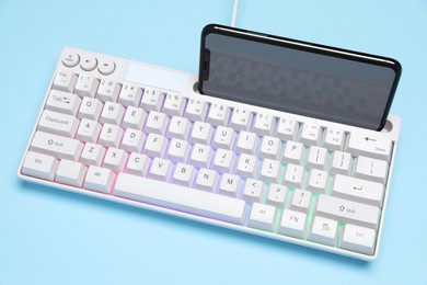 Photo of Modern white keyboard with RGB lighting and smartphone on light blue background, above view