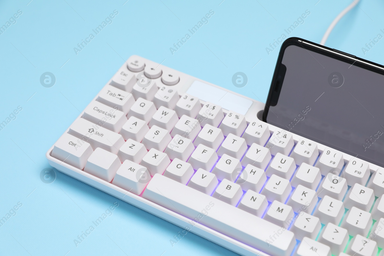 Photo of Modern white keyboard with RGB lighting and smartphone on light blue background, closeup