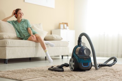 Photo of Woman in headphones resting on sofa at home, focus on vacuum cleaner