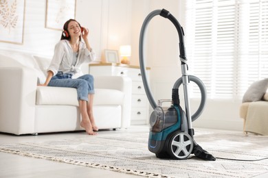 Photo of Woman in headphones resting on sofa at home, focus on vacuum cleaner