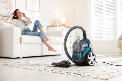 Photo of Woman in headphones resting on sofa at home, focus on vacuum cleaner