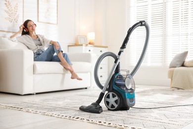 Photo of Woman in headphones resting on sofa at home, focus on vacuum cleaner
