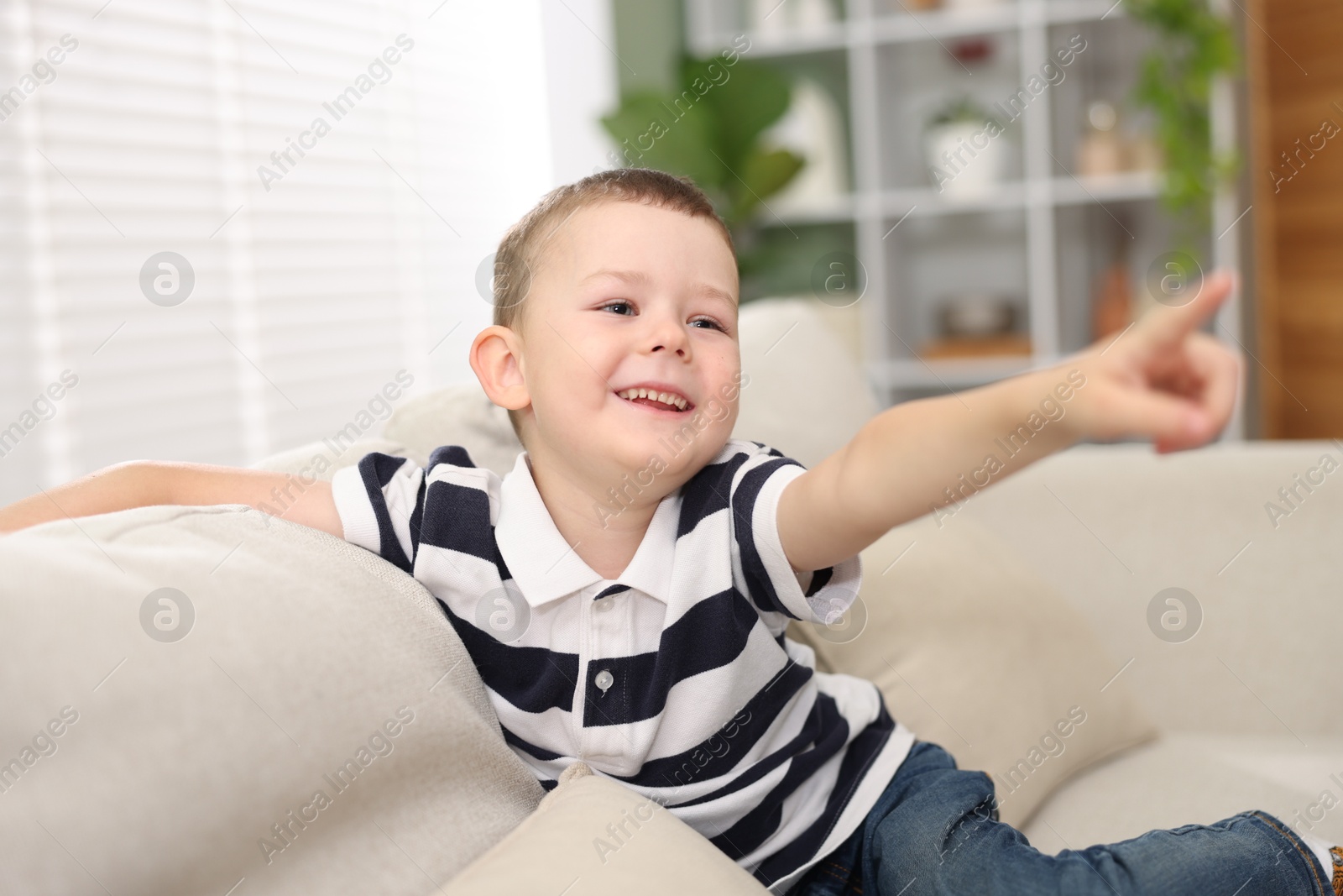 Cute little boy pointing and laughing at something on sofa indoors Photo of Cute little boy pointing and laughing at something on sofa indoors