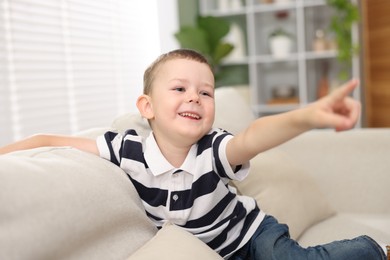 Photo of Cute little boy pointing and laughing at something on sofa indoors