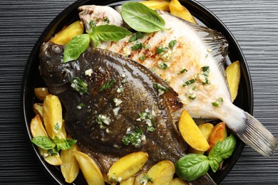 Photo of Tasty roasted flounder fish with herbs and potato on black table, top view. Homemade seafood dish