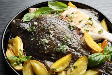 Photo of Tasty roasted flounder fish with herbs and potato on black table, closeup. Homemade seafood dish