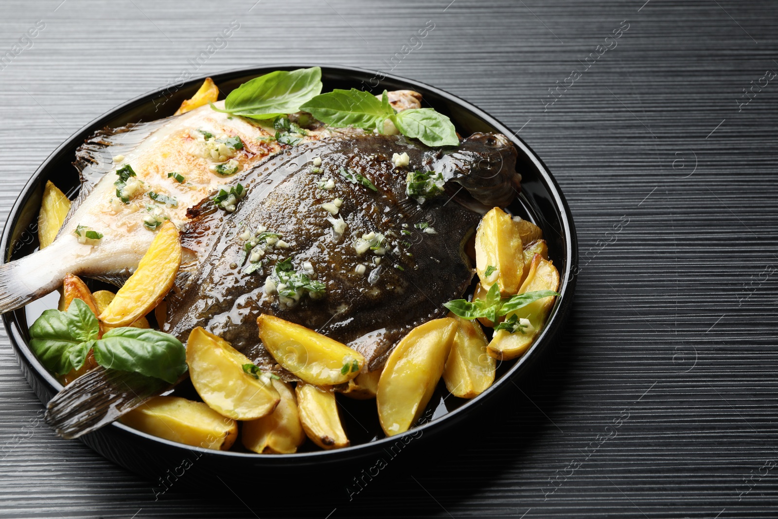 Photo of Tasty roasted flounder fish with herbs and potato on black table, closeup. Homemade seafood dish
