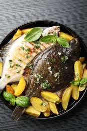 Photo of Tasty roasted flounder fish with herbs and potato on black table, top view. Homemade seafood dish