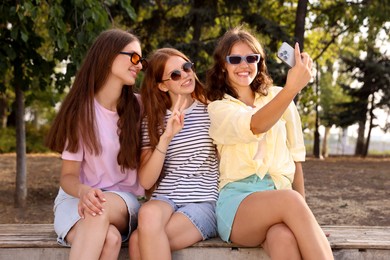 Photo of Happy friends in sunglasses taking selfie on bench outdoors