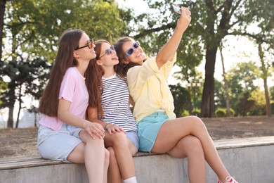Photo of Happy friends in sunglasses taking selfie on bench outdoors