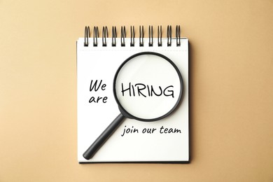 Image of We are hiring, join our team. Notebook with inscription and magnifying glass on beige background, top view