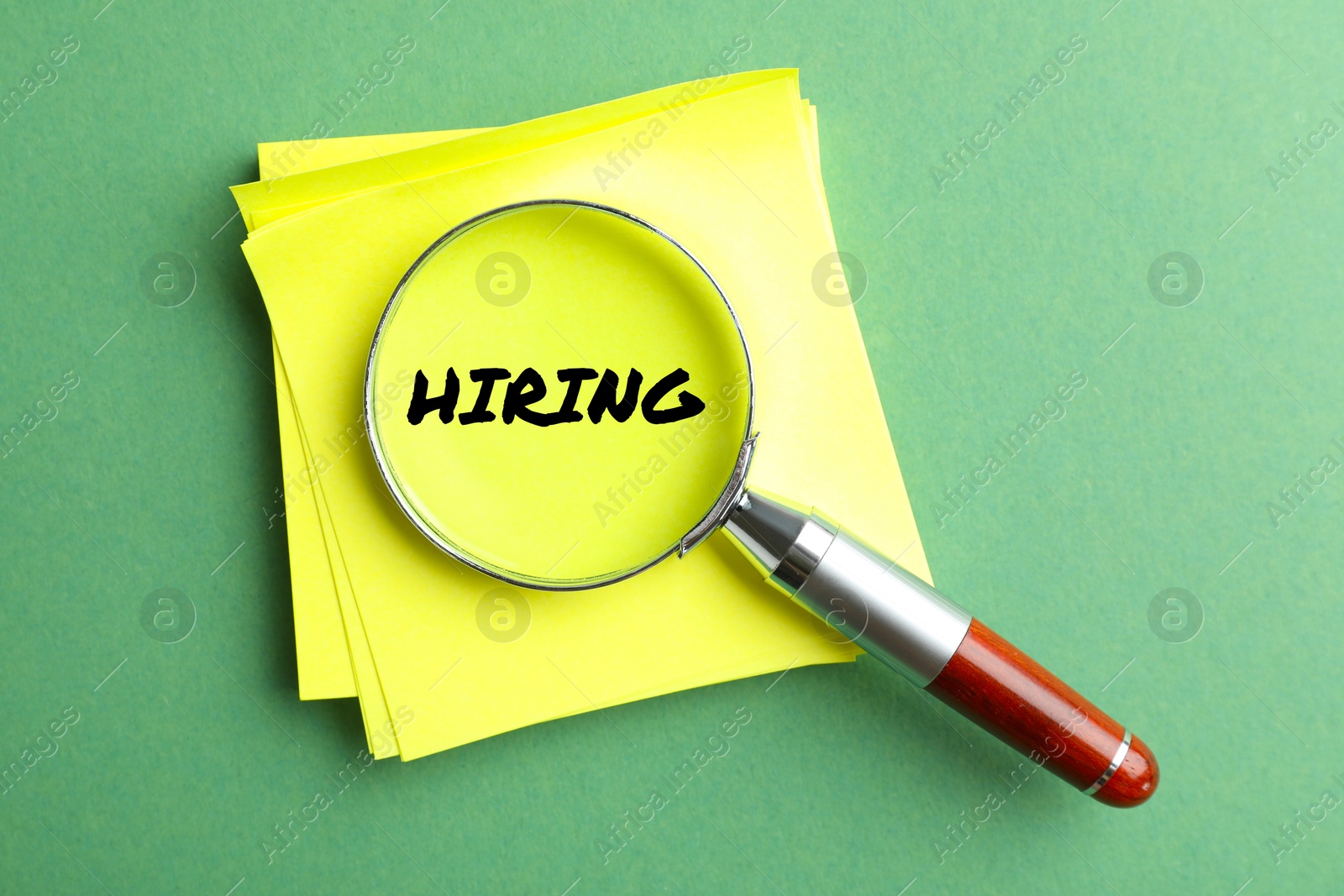 Image of Magnifying glass on note paper with word Hiring and on green background, top view.