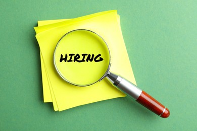 Image of Magnifying glass on note paper with word Hiring and on green background, top view.