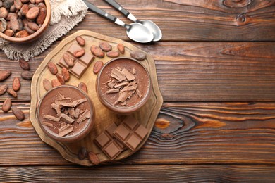 Photo of Delicious chocolate panna cotta in glasses, cocoa beans and chocolate pieces on wooden table, flat lay. Space for text