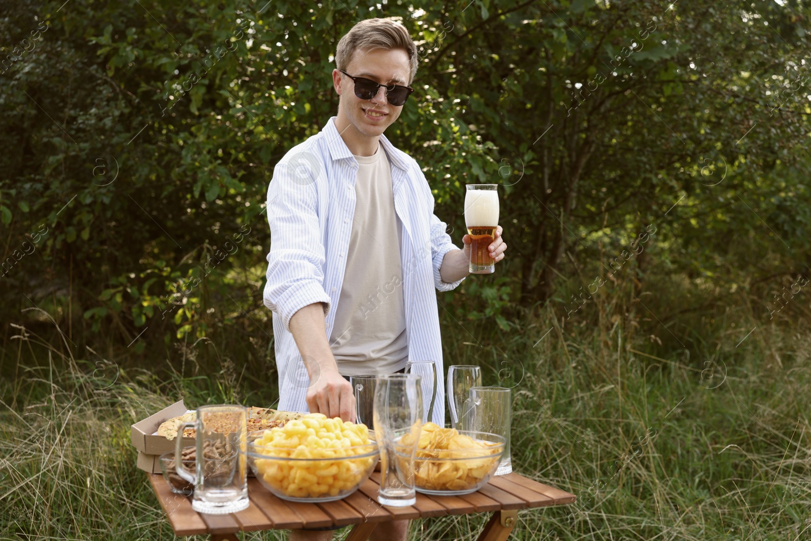 Photo of Young man holding glass of beer near wooden table with snacks outdoors