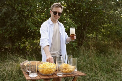 Photo of Young man holding glass of beer near wooden table with snacks outdoors