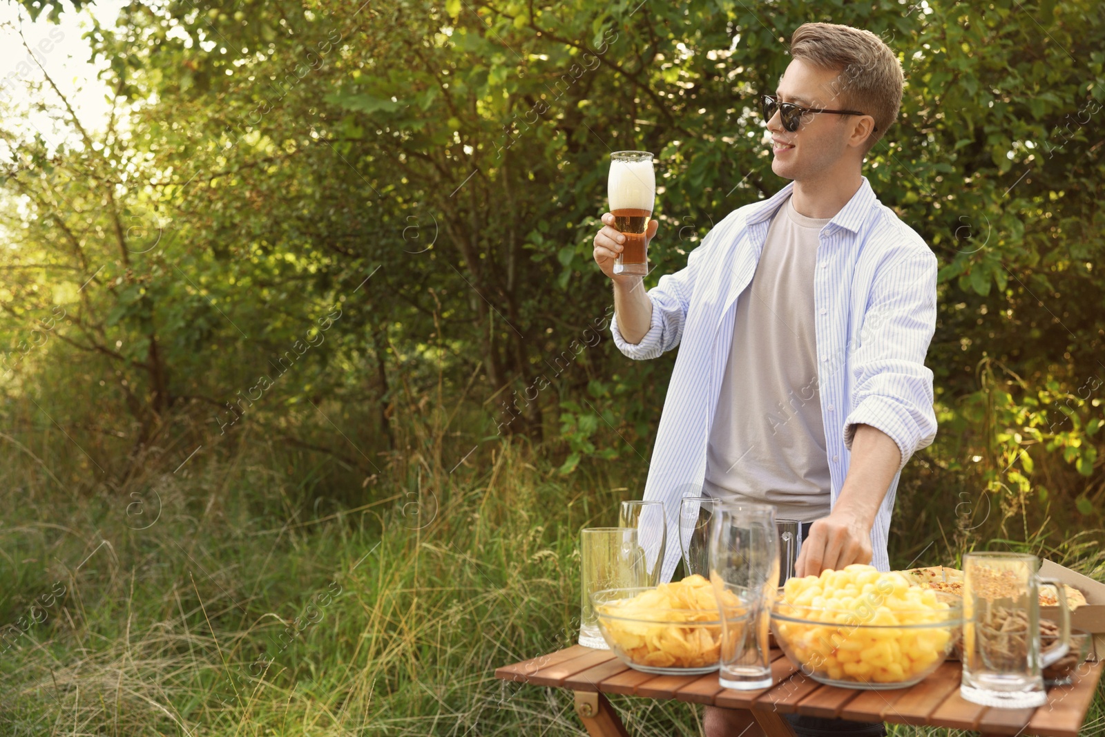 Photo of Young man holding glass of beer near wooden table with snacks outdoors, space for text