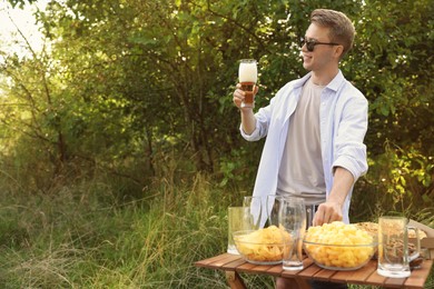 Photo of Young man holding glass of beer near wooden table with snacks outdoors, space for text