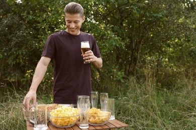 Photo of Young man holding glass of beer near wooden table with snacks outdoors, space for text