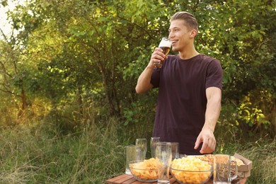 Photo of Young man holding glass of beer near wooden table with snacks outdoors, space for text