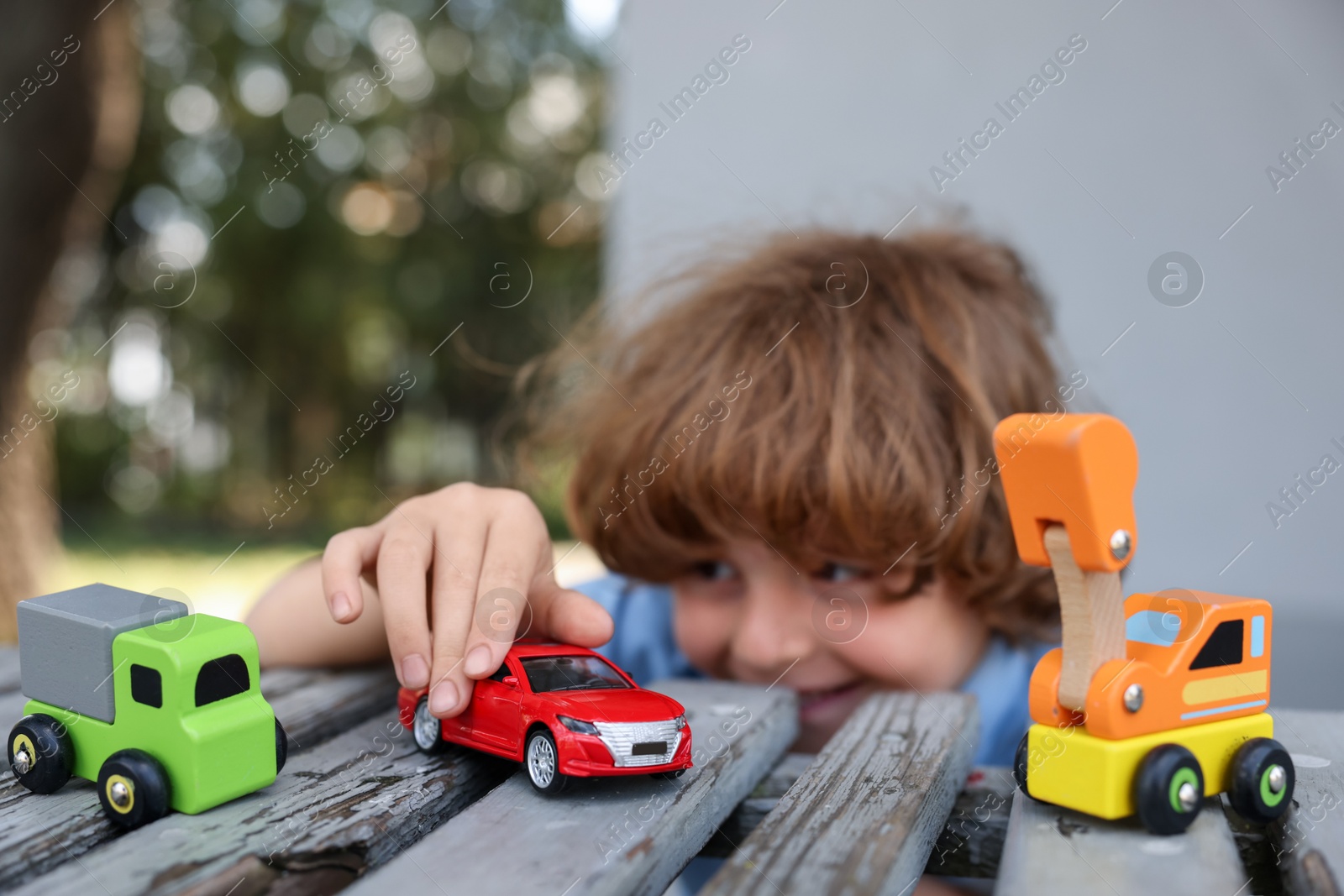 Little boy playing with toy cars on wooden bench outdoors, selective focus Photo of Little boy playing with toy cars on wooden bench outdoors, selective focus