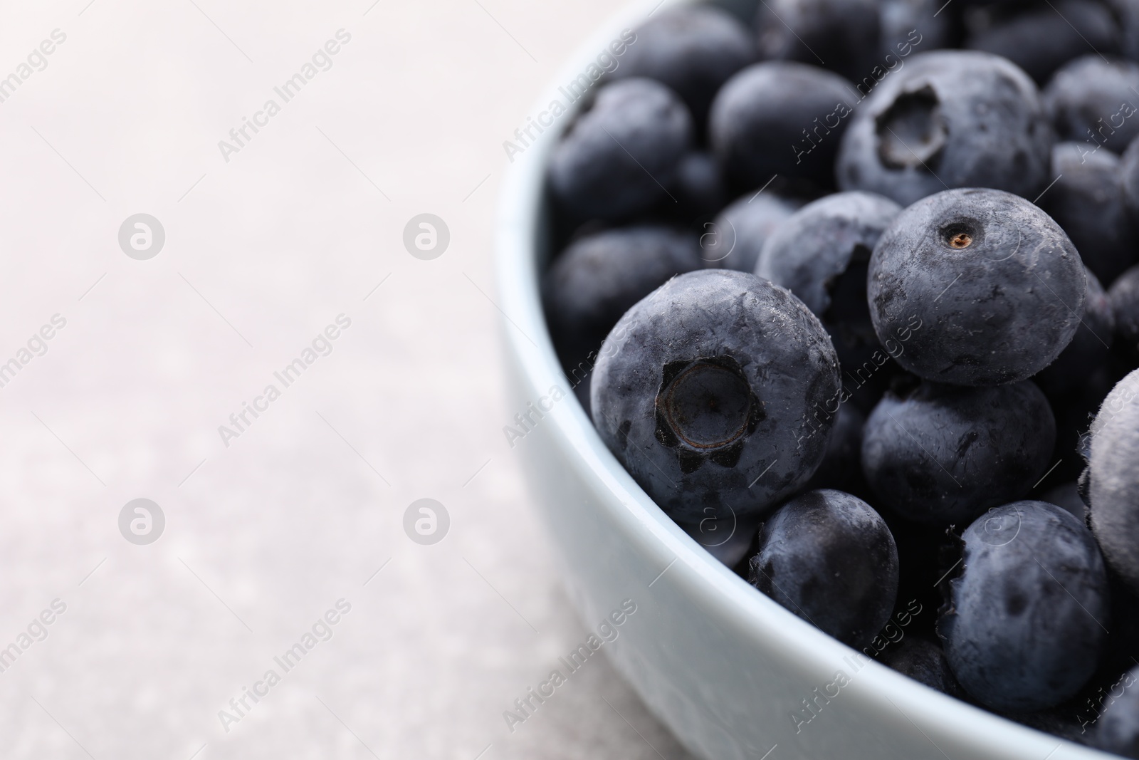 Photo of Ripe frozen blueberries in bowl on light grey table, closeup. Space for text