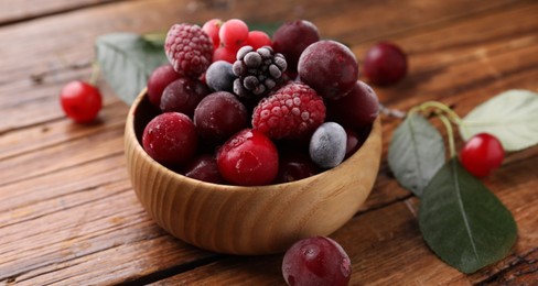 Photo of Mix of ripe frozen berries in bowl and green leaves on wooden table, closeup